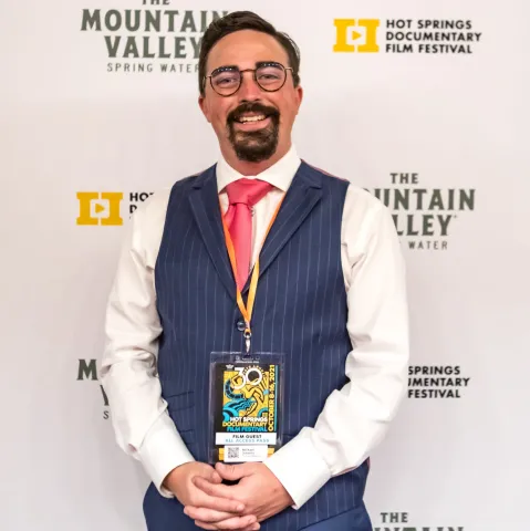 Man with a goatee wearing white long sleeved shirt with a blue vest and a red tie