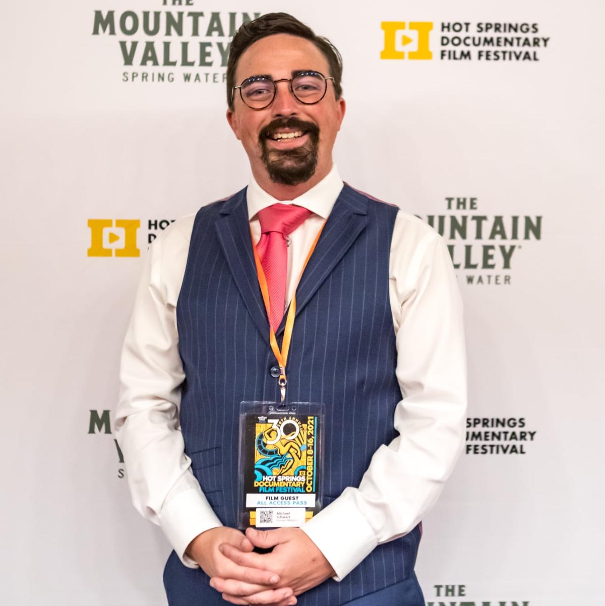 Man with a goatee wearing white long sleeved shirt with a blue vest and a red tie